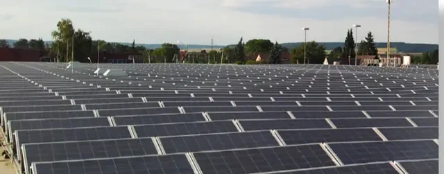 Luftaufnahme eines Flachdachs: reihenweise aufgeständerte Photovoltaikmodule reichen bis zum Horizont, dahinter ein Ortsrand mit Hügelkette.